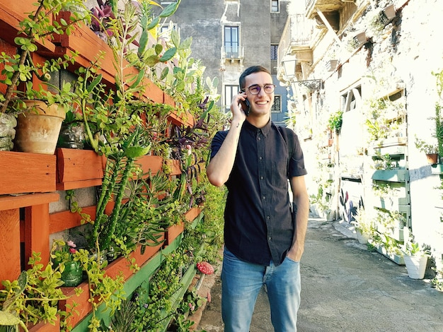 Photo young man talking on mobile phone while standing against potted plants
