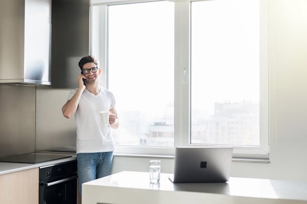 Young man talking on the phone and drinking coffee or tea while standing in the kitchen at home