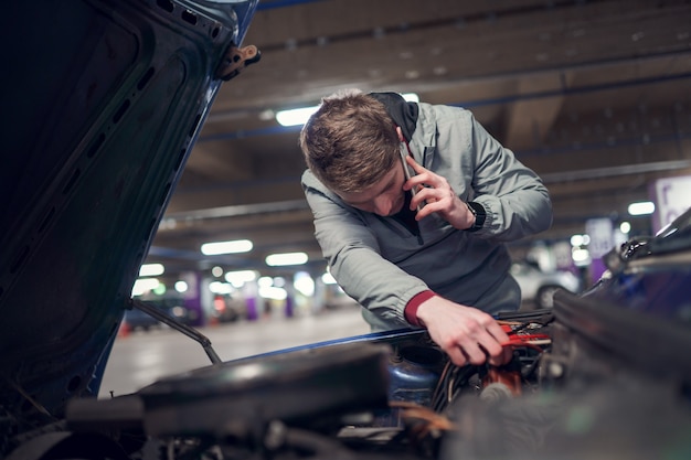 Young man talking on phone fixing car with open hood