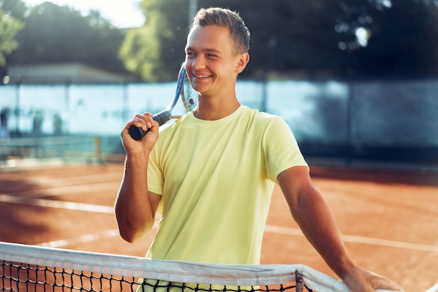 Young man teenager with tennis racket standing near net on clay tennis court