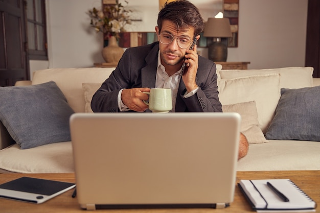 Young man teleworking from home in video conference, sitting on the couch in a suit and shorts. Drink coffee and talk on the cell phone.