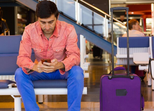 Young man texting on cellphone sitting by carry on luggage at airport terminal