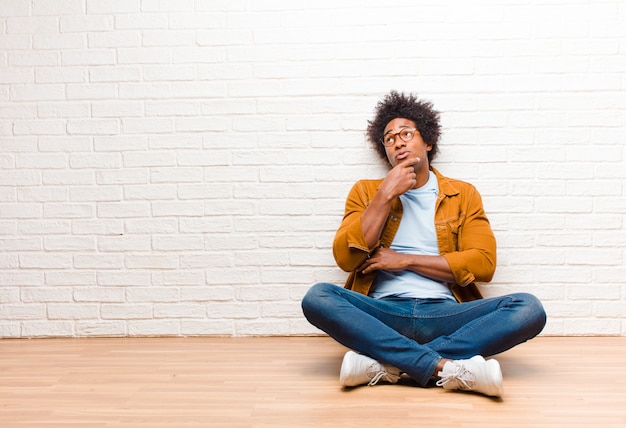 Young man thinking, feeling doubtful and confused, with different options, wondering which decision to make sitting on the floor at home