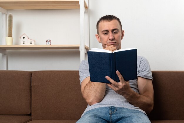 Photo young man thoughtfully reads book while sitting on sofa in living room.