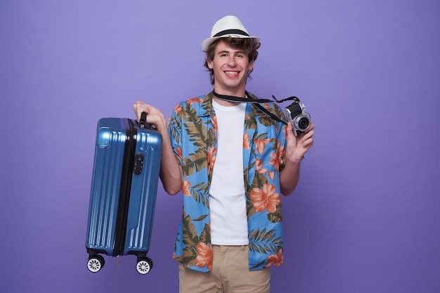 Young man tourist with luggage and camera isolated on purple studio background