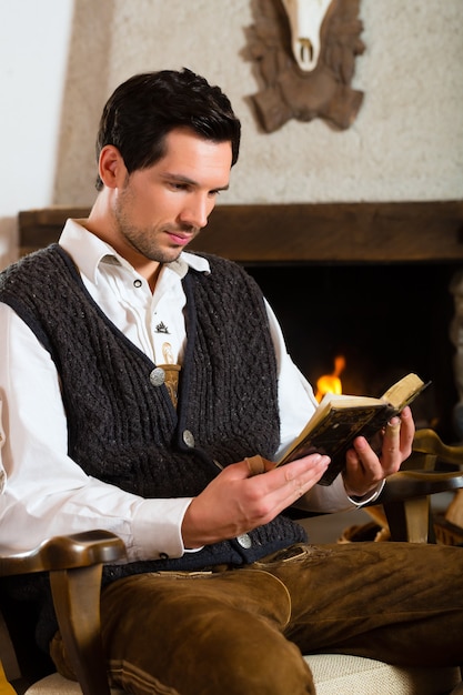 Молодой человек в традиционной горной хижине с камином читает библию