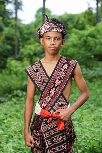 A young man in a traditional outfit stands in a field with trees in the background.