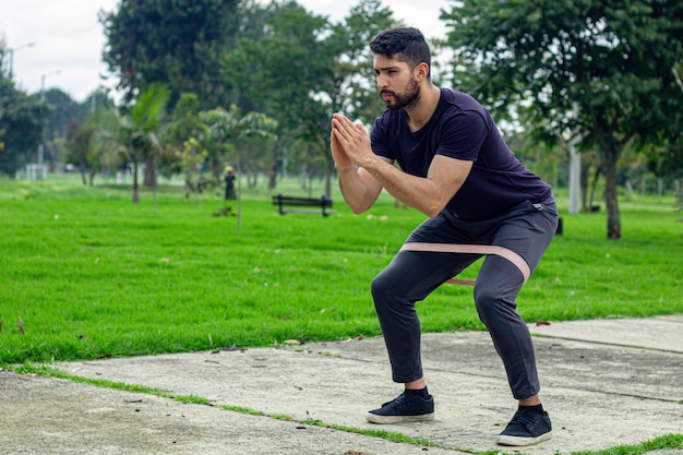 Молодой человек тренируется с эластичными полосками, занимается упражнениями на ногах на открытом воздухе в парке.