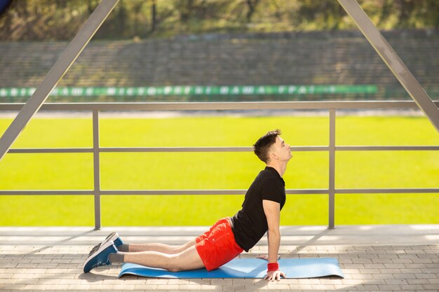 若い男が屋外でヨガをトレーニングします。スポーティな男は、スポーツグラウンドで、青いヨガマットでストレッチ運動を行います。