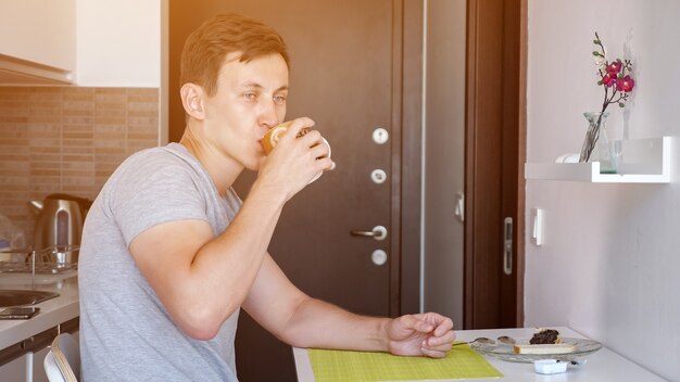 Young man traveler is having a breakfast. He is sitting at table and drinking tea, side view. He is in small kitchen in rented flat on vacation, sunlight