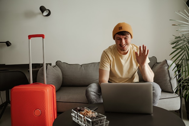 Young man traveler is sitting on couch and speaking with someone on stream in hotel room on summer vacation. Cool male in casual clothing with luggage.