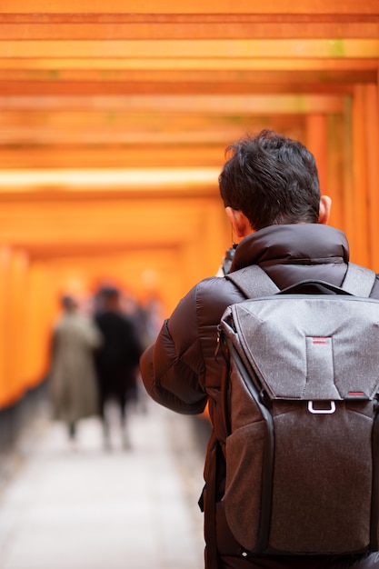 Молодой человек путешествуя на святыне fushimi inari taisha, счастливый азиатский путешественник смотря яркие оранжевые ворота torii. достопримечательности и популярные для туристов достопримечательности в киото, япония. концепция путешествия по азии