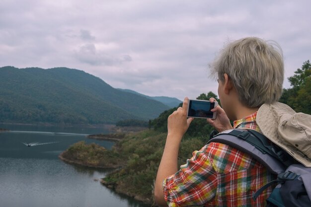 若い男のトレッカーを使用してスマートフォンを取る写真は、バックパックで旅行を楽しむ