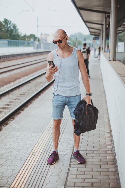 Молодой человек в футболке на платформе ждет поезд с помощью мобильного телефона на вокзале pl