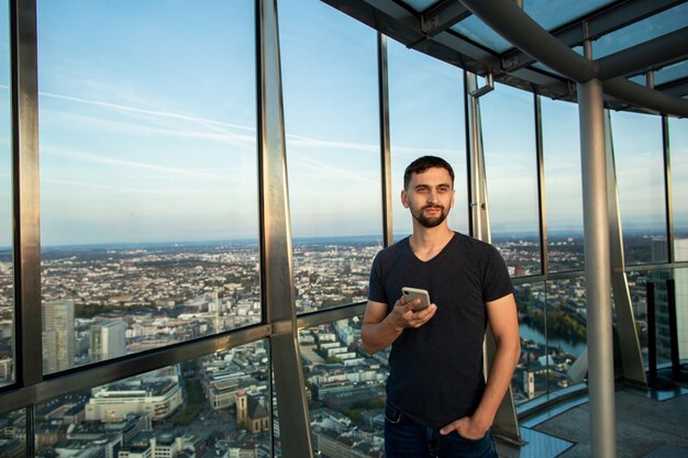 Young man in tshirt stands on the observation deck, holding cellphone and typing text message.