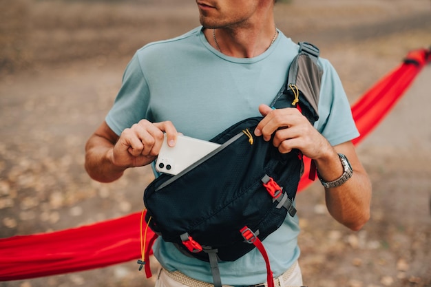 A young man in a tshirt and a waist bag on his shoulder puts a\
smartphone in his pocket close up