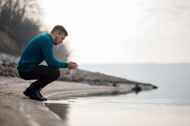 A young man in a turquoise sweater wets his hands in the river