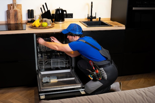 Young man in uniform with a set of tools repairs a dishwasher in the kitchen