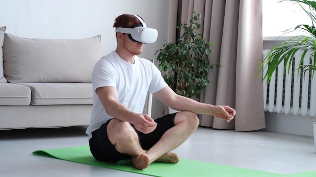 Young man using AR while meditating sitting in lotus position on roll mat wearing sports wear indoors man wearing virtual reality device