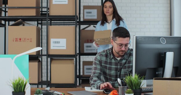 A young man using a computer a woman confirms the contact details on the boxes A man and a woman work in a warehouse