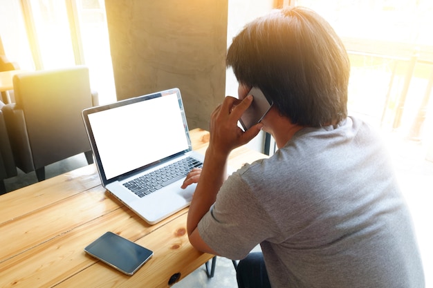 Giovane uomo utilizzando computer portatile con schermo in bianco spazio copia e chiamando cellulare mentre seduto in caffè, sole flare