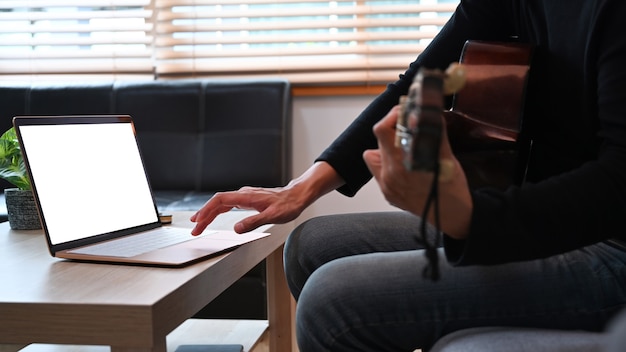 Young man using laptop and playing guitar in living room.