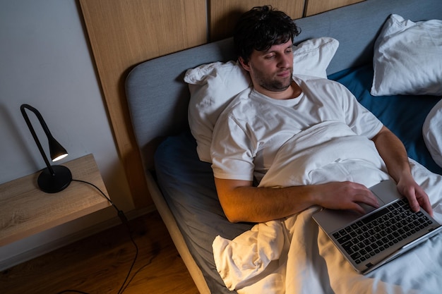 A young man using a laptop while lying on a bed at night Man is resting in bed with a movie on his laptop