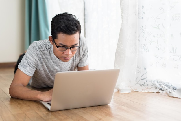 young man using laptop while lying on floor by window at home