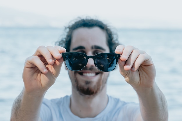 Молодой человек, использующий солнцезащитные очки, много улыбаясь, прикасаясь к ним, концепция солнцезащитных очков, лето и путешествия, копия пространства