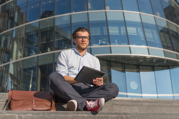 Young man using tablet pc sitting on stairs near modern glass building