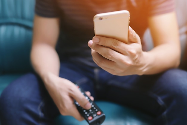 Young man using television remote control and mobile smart phone  is sitting on a sofa. on or off tv.