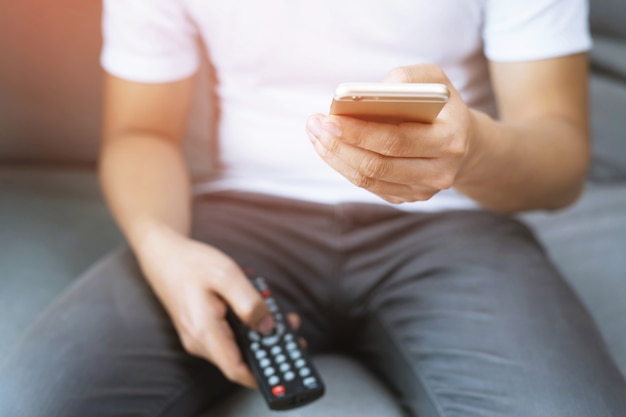 Young man using television remote control and mobile smart phone  is sitting on a sofa