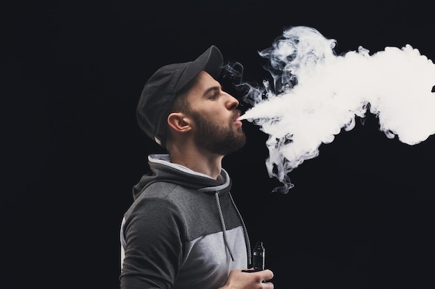 Young man vaping, studio shot. Bearded brutal guy in cap blowing a cloud of smoke on black background. Nicotine free smoking and vapor concept, copy space