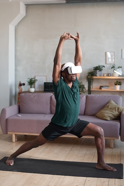 Young man in VR glasses standing on exercise mat and exercising in the room