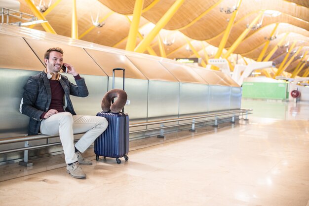 Young man waiting listening music and using mobile phone at the airport with a suitcase.