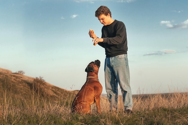 A young man on a walk eats a fast food hamburger and trains his\
dog