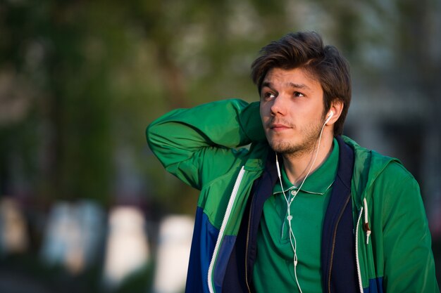 Young man walking around city listening to music with ear headphones looks and smiling. Close-up shot. Sunset. Autumn weather.