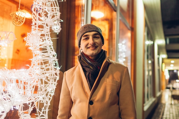 Young man walking in city by decorated Christmas showcases at night and looking at camera.