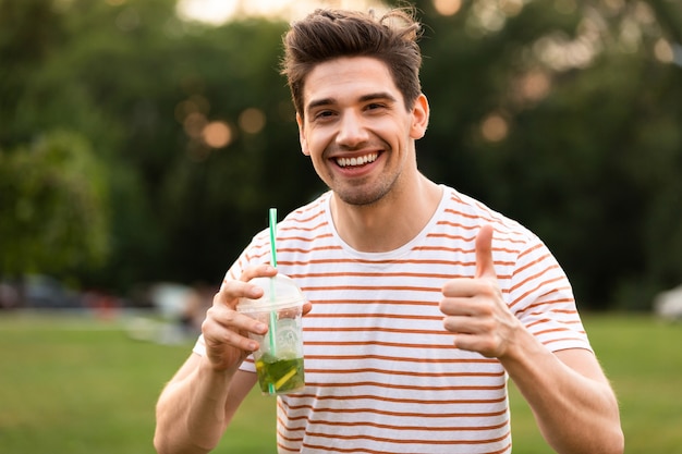 緑の公園を歩いて、プラスチック製のコップから飲み物を飲む若い男