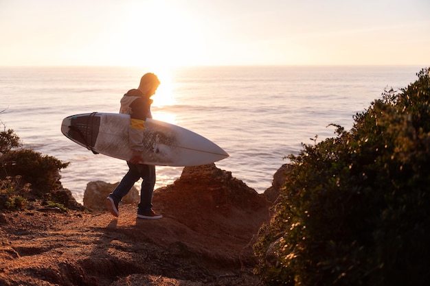 Молодой человек, идущий на закате со своей доской для серфинга вдоль скал у моря, концепция досуга и хобби копирует пространство для текста