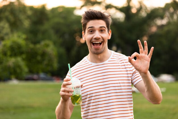 公園を歩いて、プラスチック製のコップから飲み物を飲む若い男