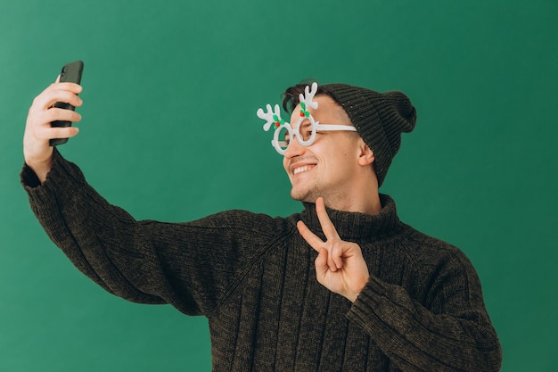 따뜻한 스웨터 카니발 안경을 쓴 청년과 녹색 배경에 고립된 손에 전화기