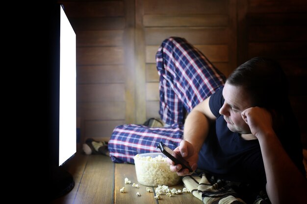 Молодой человек смотрит телевизор дома на полу и ест попкорн