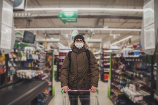 Young man in wear casual winter clothes with glasses and medical mask Ñ grocery cart in hypermarket. Attractive teenager with backpack in city shop, looking at camera and emotions of an unexpected