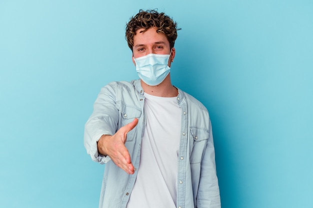 Young man wearing an antiviral mask isolated on blue wall stretching hand at camera in greeting gesture