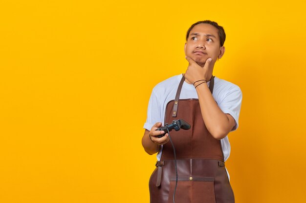 Young man wearing apron holding game controller looking aside with doubtful and skeptical expression