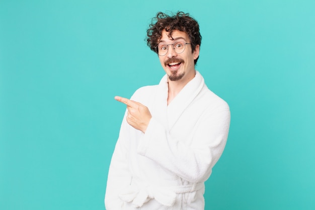 Young man wearing bathrobe looking excited and surprised pointing to the side