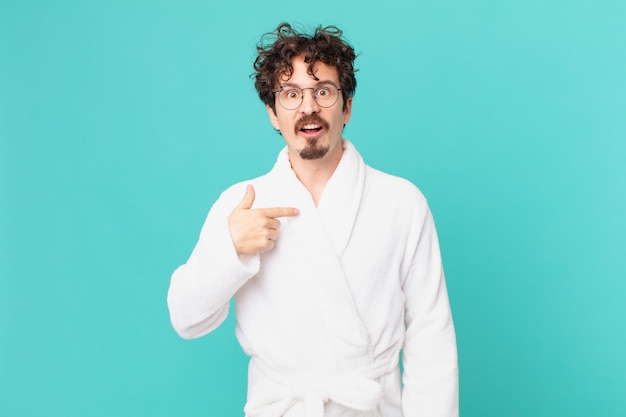 Young man wearing bathrobe looking shocked and surprised with mouth wide open, pointing to self