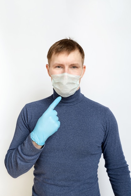Young man wearing blue medical rubber gloves and medical mask points at his face on white background
