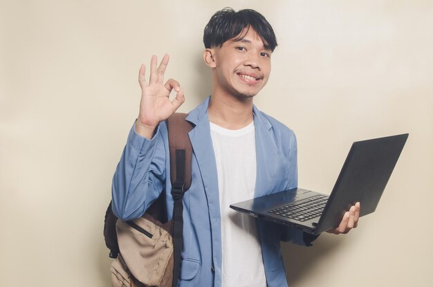 Young man wearing college suit with OK gesture while carrying laptop on isolated background